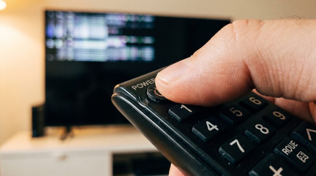 Mão apertando o botão de ligar do controle remoto enquanto a televisão apresenta problemas e desliga ao fundo.