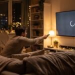 Pessoa sentada no sofá com o controle remoto na mão tentando acessar aplicativo na Smart TV que exibe tela de carregamento.