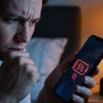 Usuário frustrado segurando o celular no escuro mostrando alerta de bateria fraca de 5%.