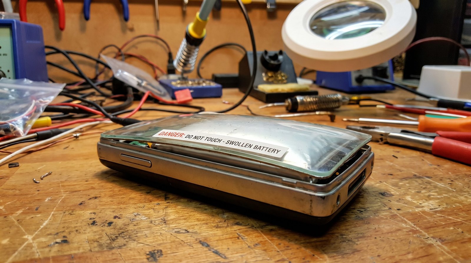 Celular antigo em uma bancada de manutenção com a tampa traseira de vidro visivelmente estufada e extremamente perigosa devido a danos irreversíveis na bateria.