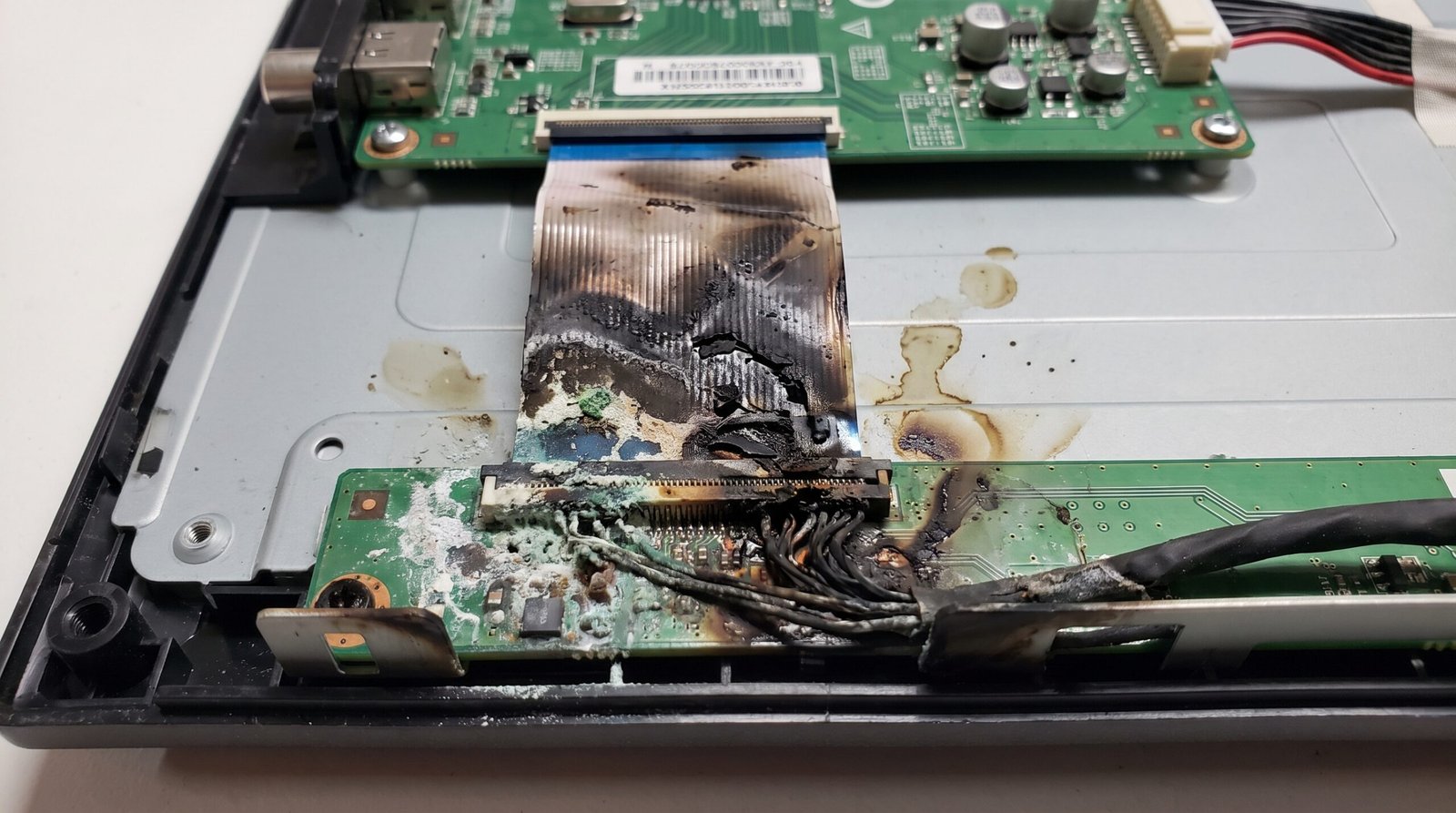 Fita de comunicação interna de uma placa de Smart TV totalmente carbonizada, queimada e oxidada na parte inferior do painel, evidenciando dano fatal e curto-circuito causado por líquidos.