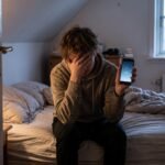 Pessoa frustrada sentada na cama do quarto distante, segurando um celular com o sinal fraco do Wi-Fi exibindo a tela de carregamento e falta de internet.
