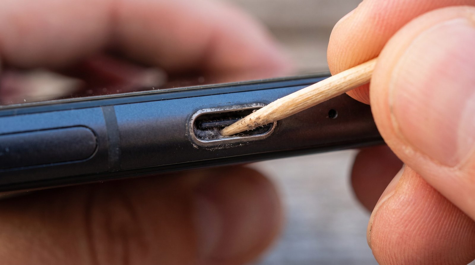 Visão detalhada de um usuário utilizando cuidadosamente um palito de madeira para limpar a sujeira e poeira acumulada dentro da porta de carregamento do celular.